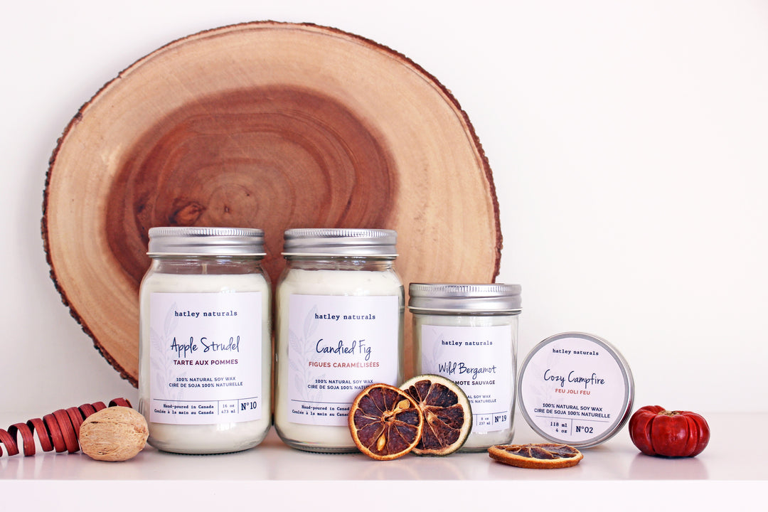 Fruit placed around and in front of 3 Candles in jars, and one candle in a tin container with a slab of a wood log in the background and a white background.